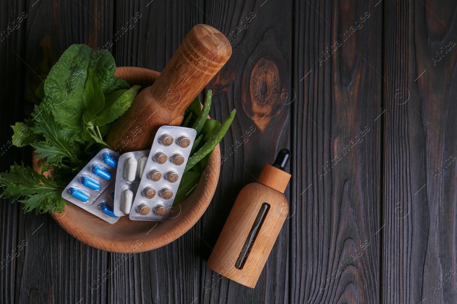 Photo of Mortar with fresh green herbs, extract and pills on wooden table, flat lay. Space for text