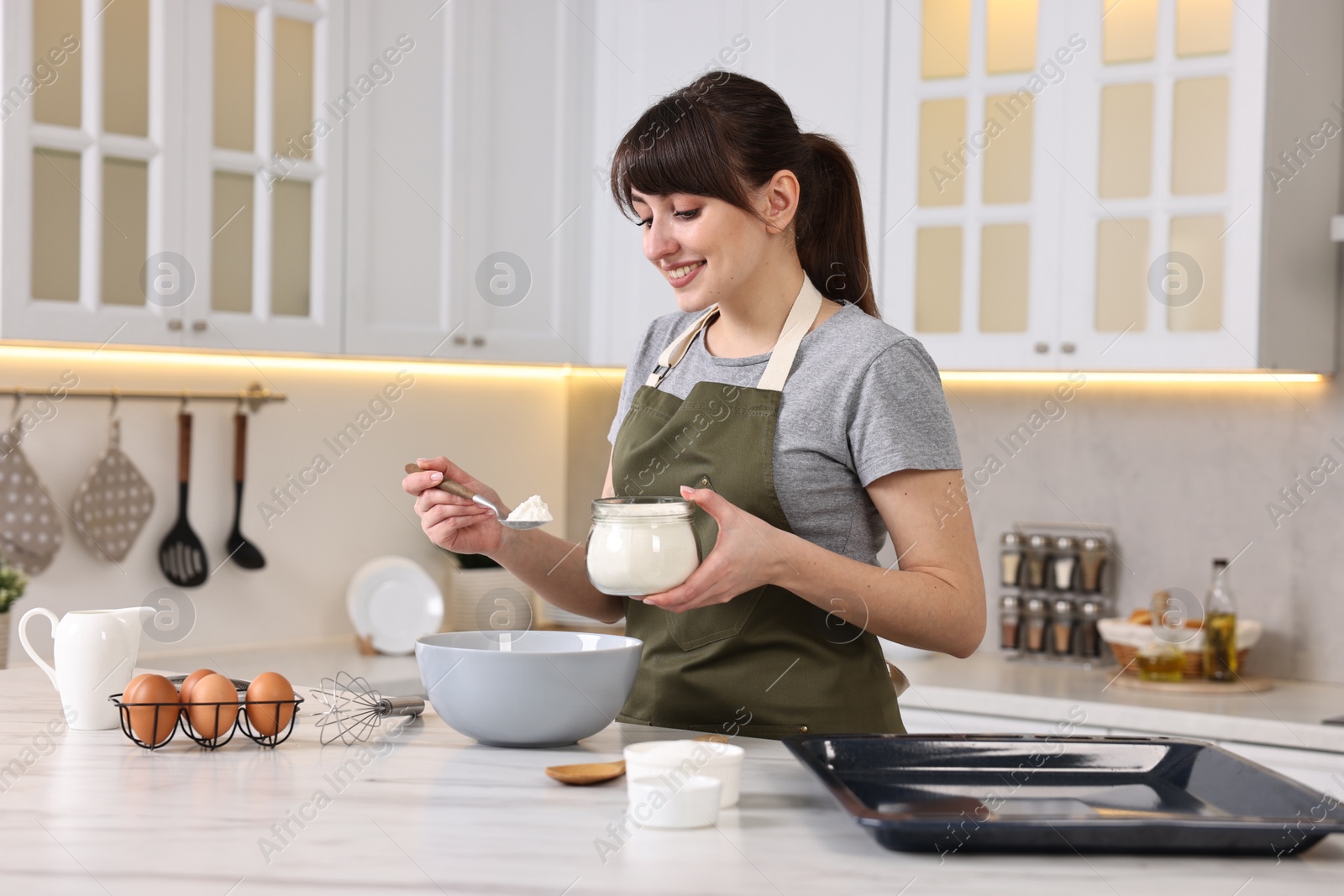 Photo of Happy young housewife adding flour into bowl at white marble table in kitchen. Cooking process