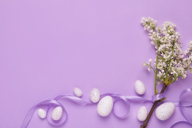 Many painted Easter eggs, branch with lilac flowers and ribbon on violet background, flat lay. Space for text