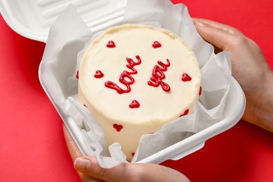 Woman holding takeaway box with bento cake at red table, closeup. St. Valentine's day surprise