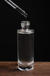 Dripping cosmetic serum from pipette into bottle at wooden table against black background