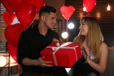 Man presenting gift to his girlfriend in room decorated with heart shaped balloons. Valentine's day celebration