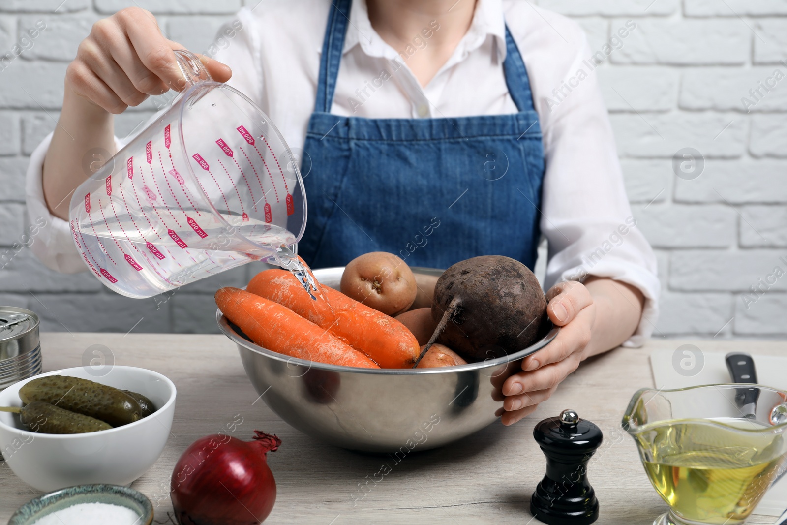 Photo of Woman pouring water into bowl with fresh vegetables at white wooden table, closeup. Cooking vinaigrette salad