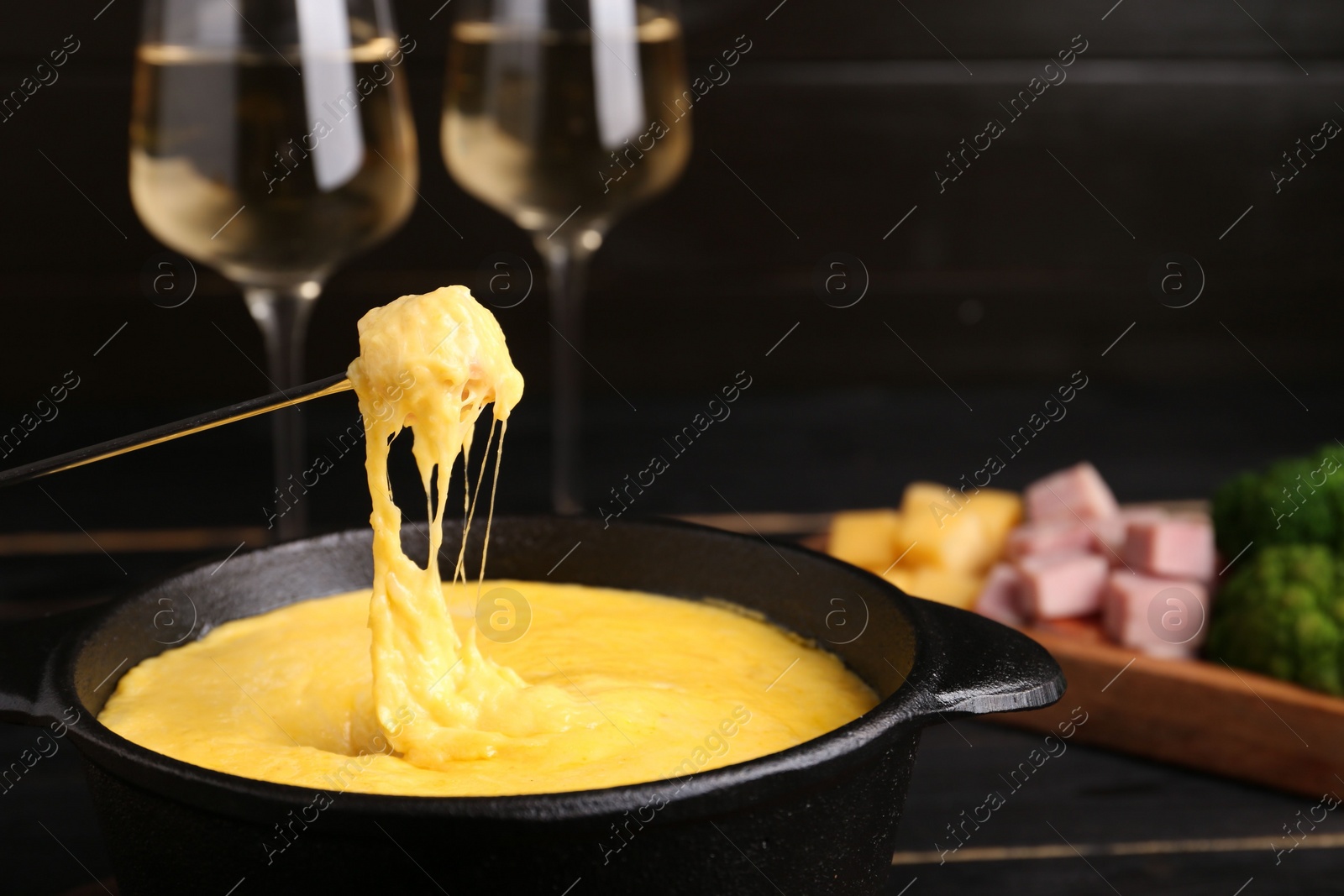 Photo of Dipping piece of ham into fondue pot with melted cheese on black wooden table, closeup