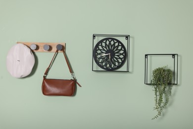 Photo of Stylish clock, houseplant and wooden rack with bag, hat on light green wall. Interior design