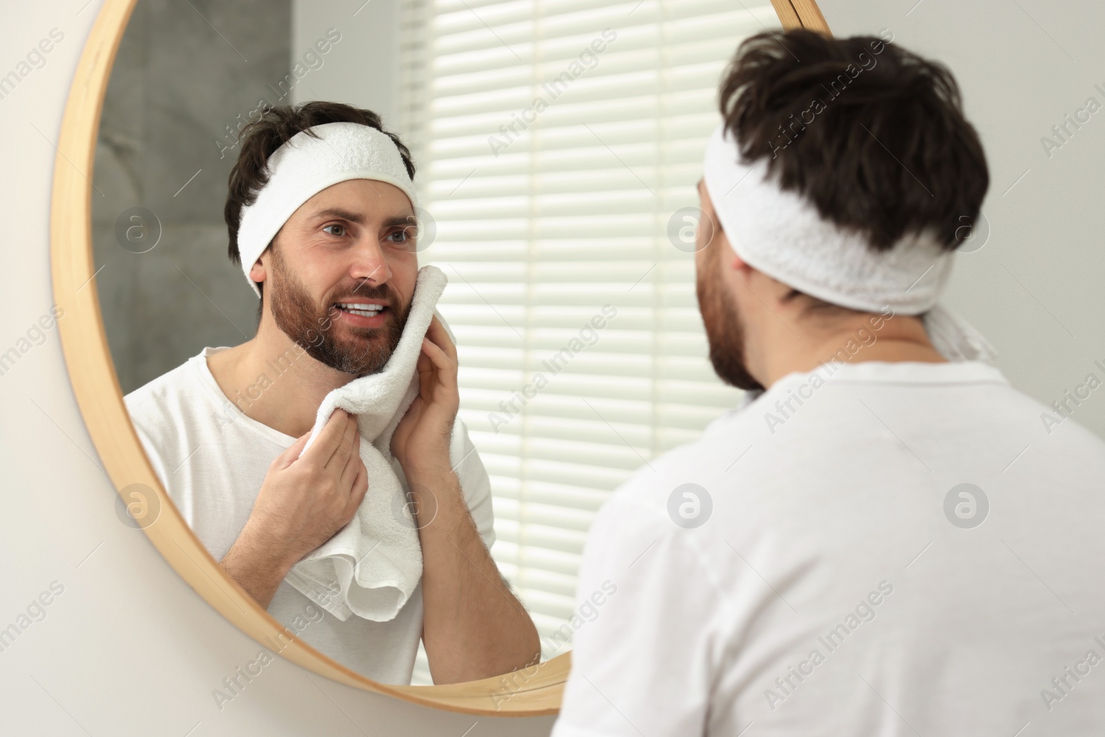 Photo of Washing face. Man with headband and towel near mirror in bathroom