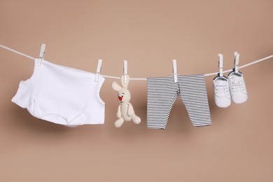 Photo of Cute small baby shoes, clothes and toy hanging on washing line against brown background