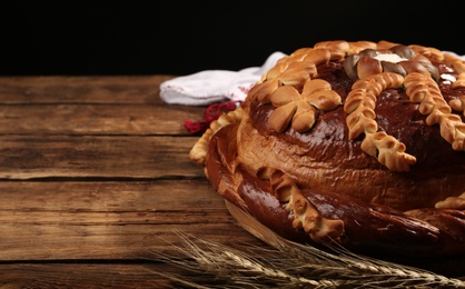 Photo of Korovai with wheat spikes on wooden table against black background, space for text. Ukrainian bread and salt welcoming tradition