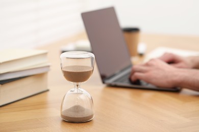 Photo of Hourglass with flowing sand on desk. Man using laptop indoors, selective focus