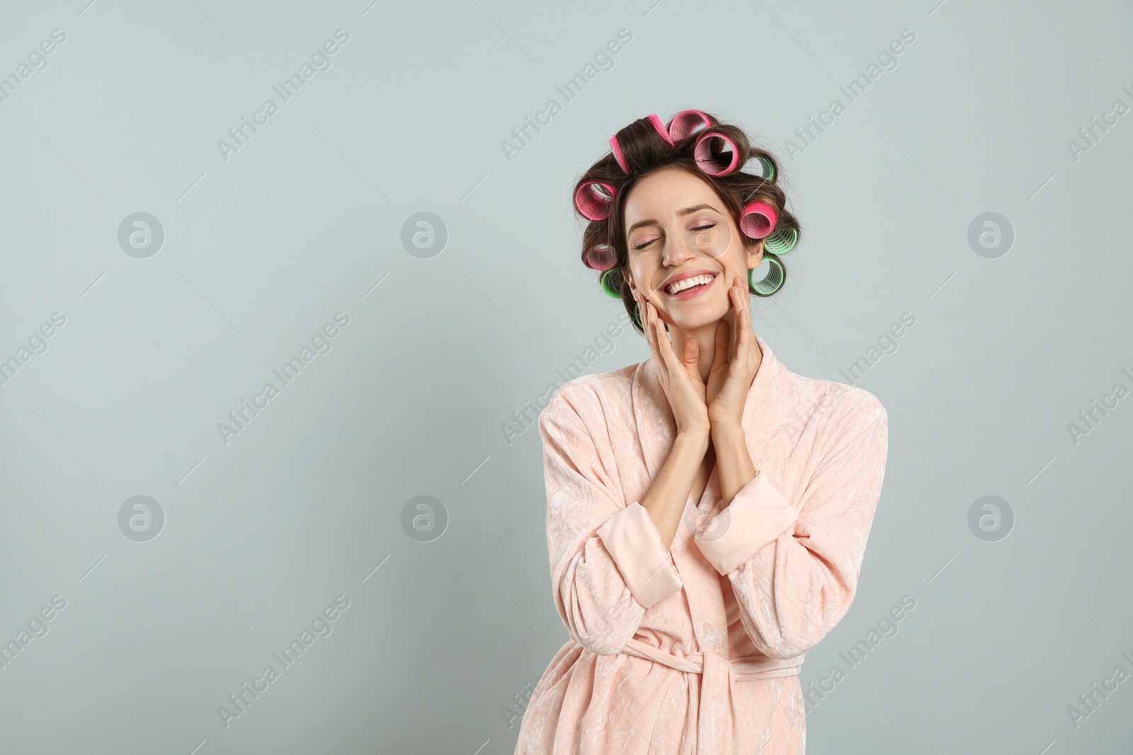 Photo of Beautiful young woman in bathrobe with hair curlers on light grey background. Space for text