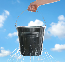 Image of Woman holding leaky bucket with water against blue sky, closeup  