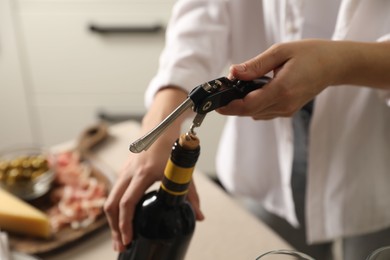 Woman opening wine bottle with corkscrew at table indoors, closeup