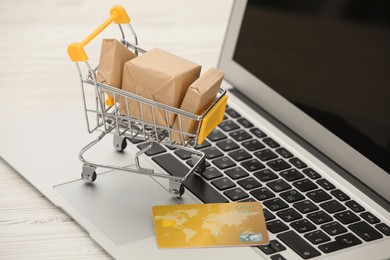 Online payment concept. Small shopping cart with bank card, boxes and laptop on white table, closeup