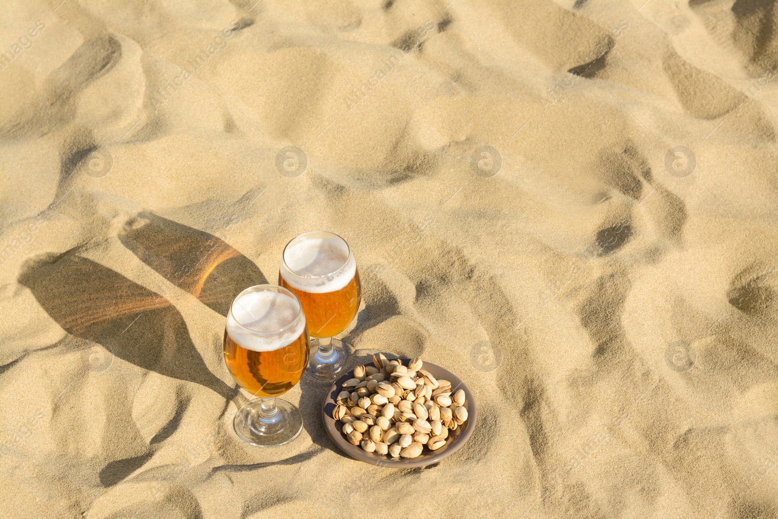 Photo of Glasses of cold beer and pistachios on sandy beach, space for text