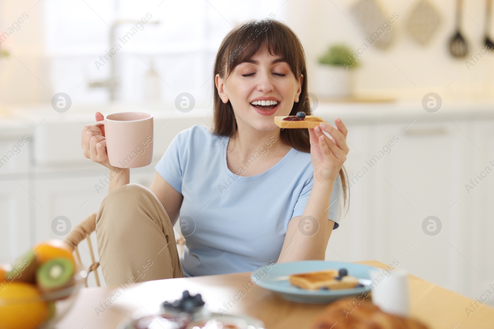Photo of Smiling woman drinking coffee and eating toast at breakfast indoors