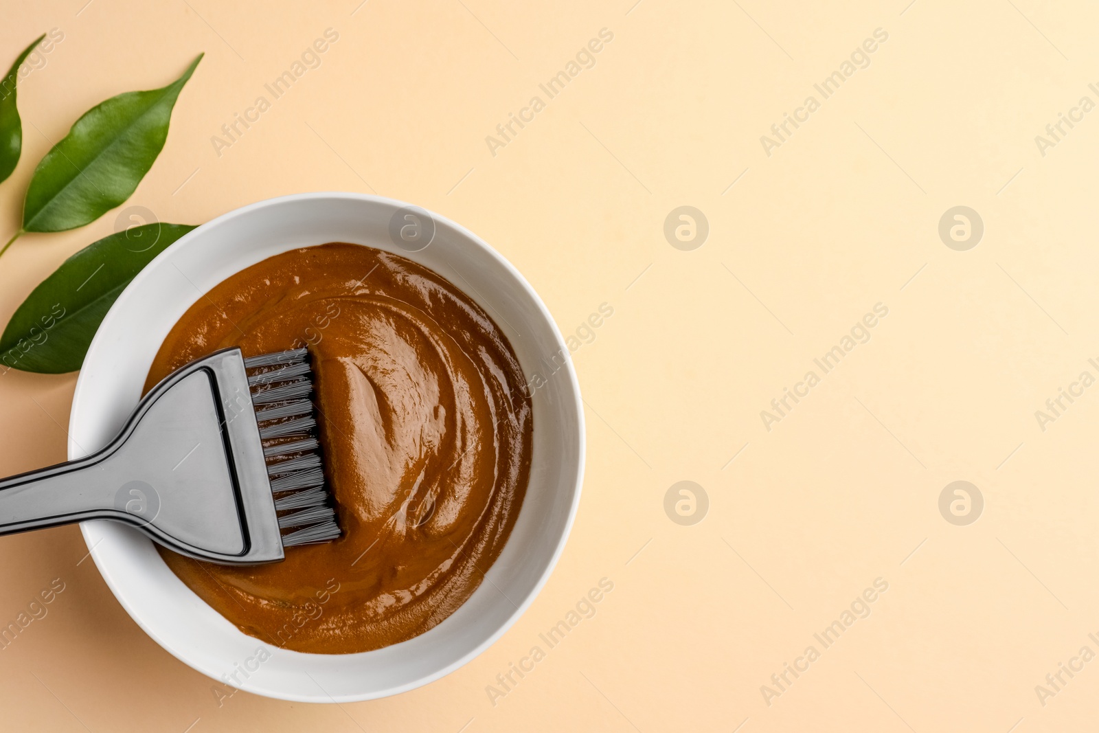 Photo of Bowl of henna cream, brush and green leaves on beige background, flat lay with space for text. Natural hair coloring