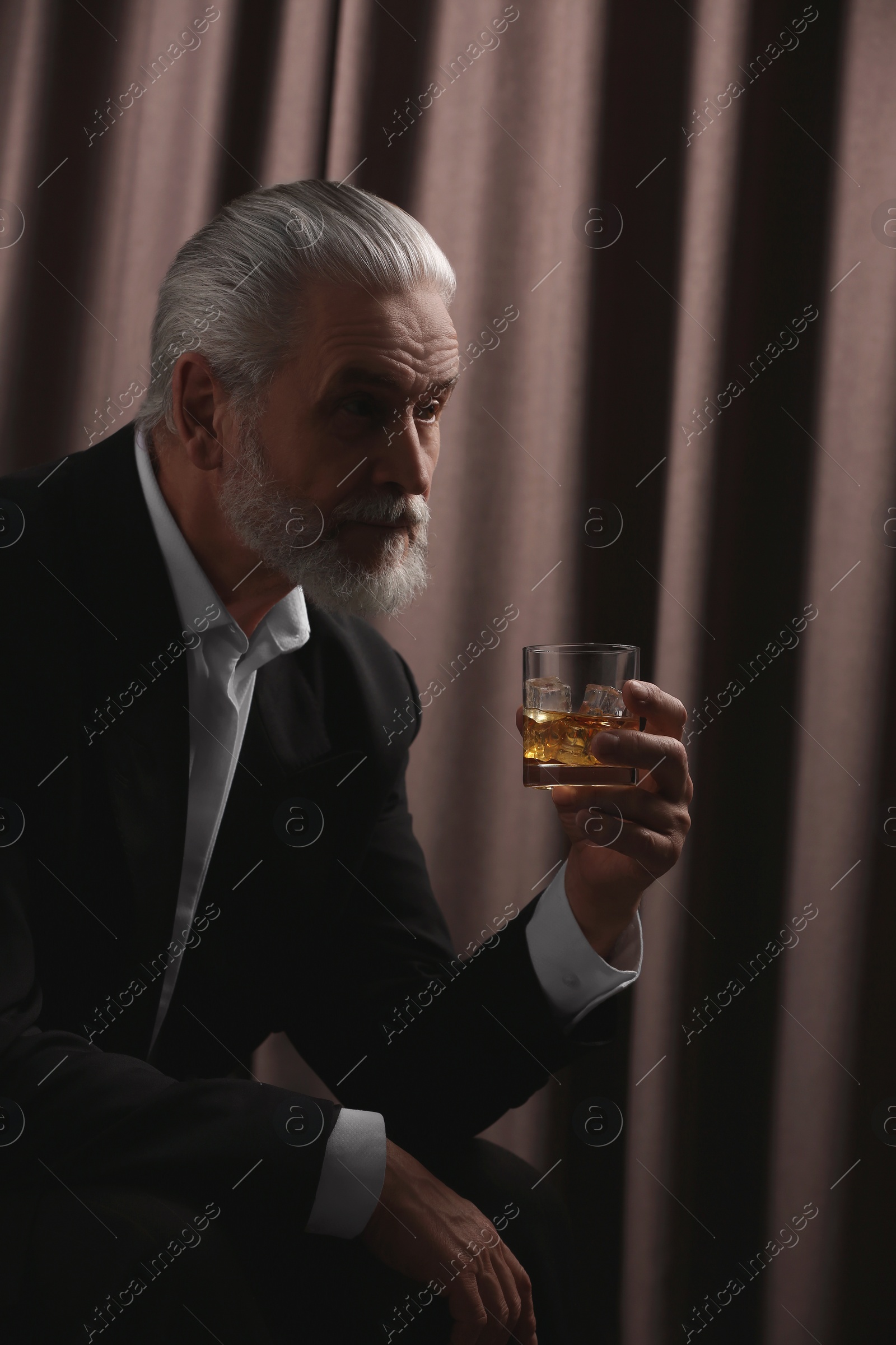 Photo of Senior man in formal suit holding glass of whiskey with ice cubes on brown background