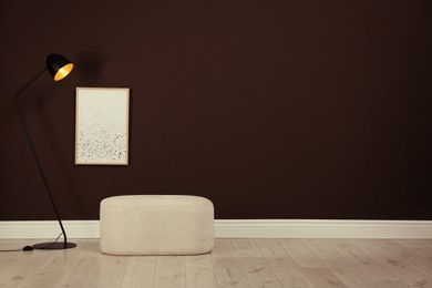Photo of Stylish ottoman and lamp near brown wall in room. Space for text