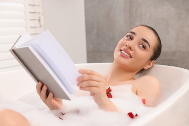 Happy woman reading book while taking bath in tub with foam and rose petals indoors
