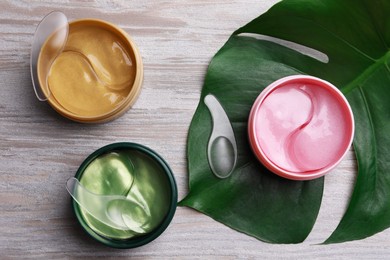 Packages of different under eye patches and tropical leaf on wooden table, flat lay. Cosmetic product