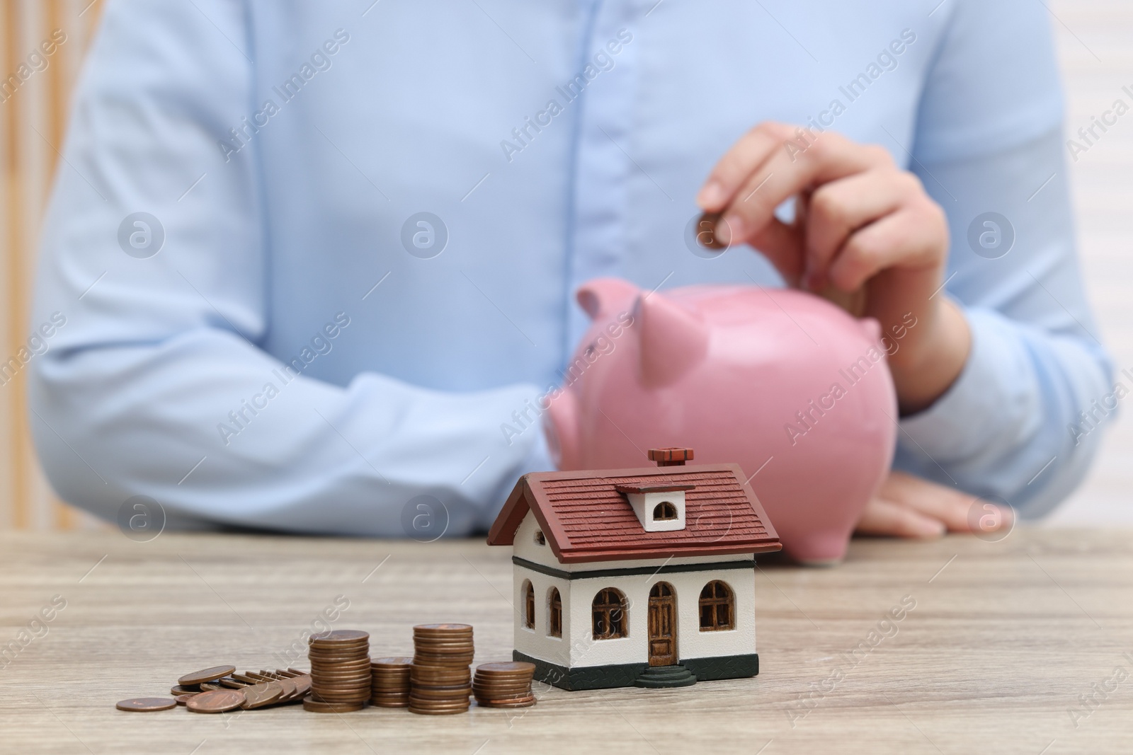 Photo of Woman with piggy bank at wooden table, focus on house model and stacked coins