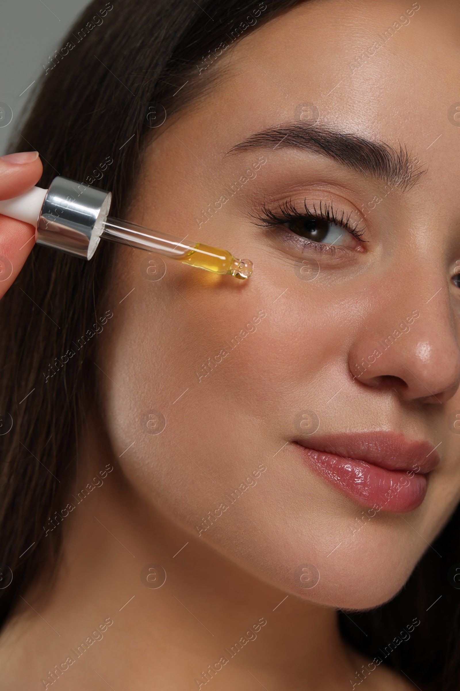 Photo of Beautiful young woman applying essential oil onto face on grey background, closeup