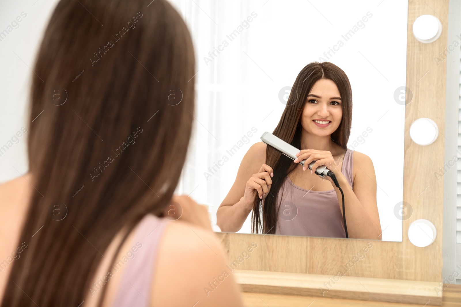 Photo of Young woman using hair iron near mirror indoors