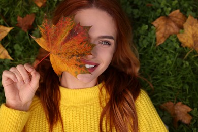 Smiling woman lying on grass and covering eye with autumn leaf, top view