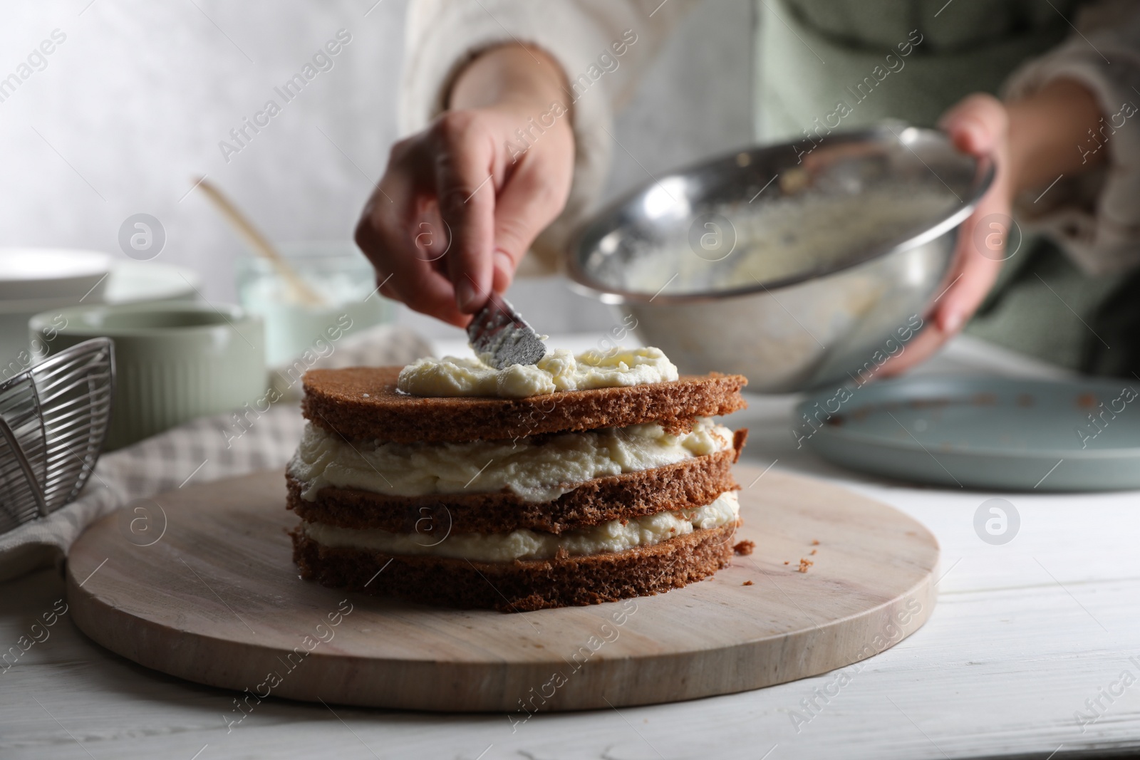 Photo of Woman smearing sponge cake with cream at white wooden table, closeup