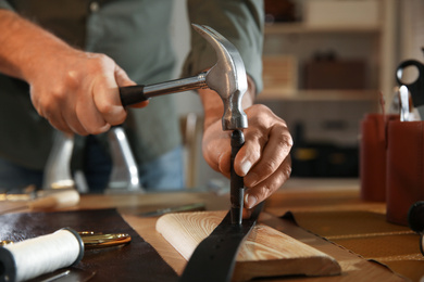 Photo of Man making holes in leather belt with punch and hammer at workshop, closeup