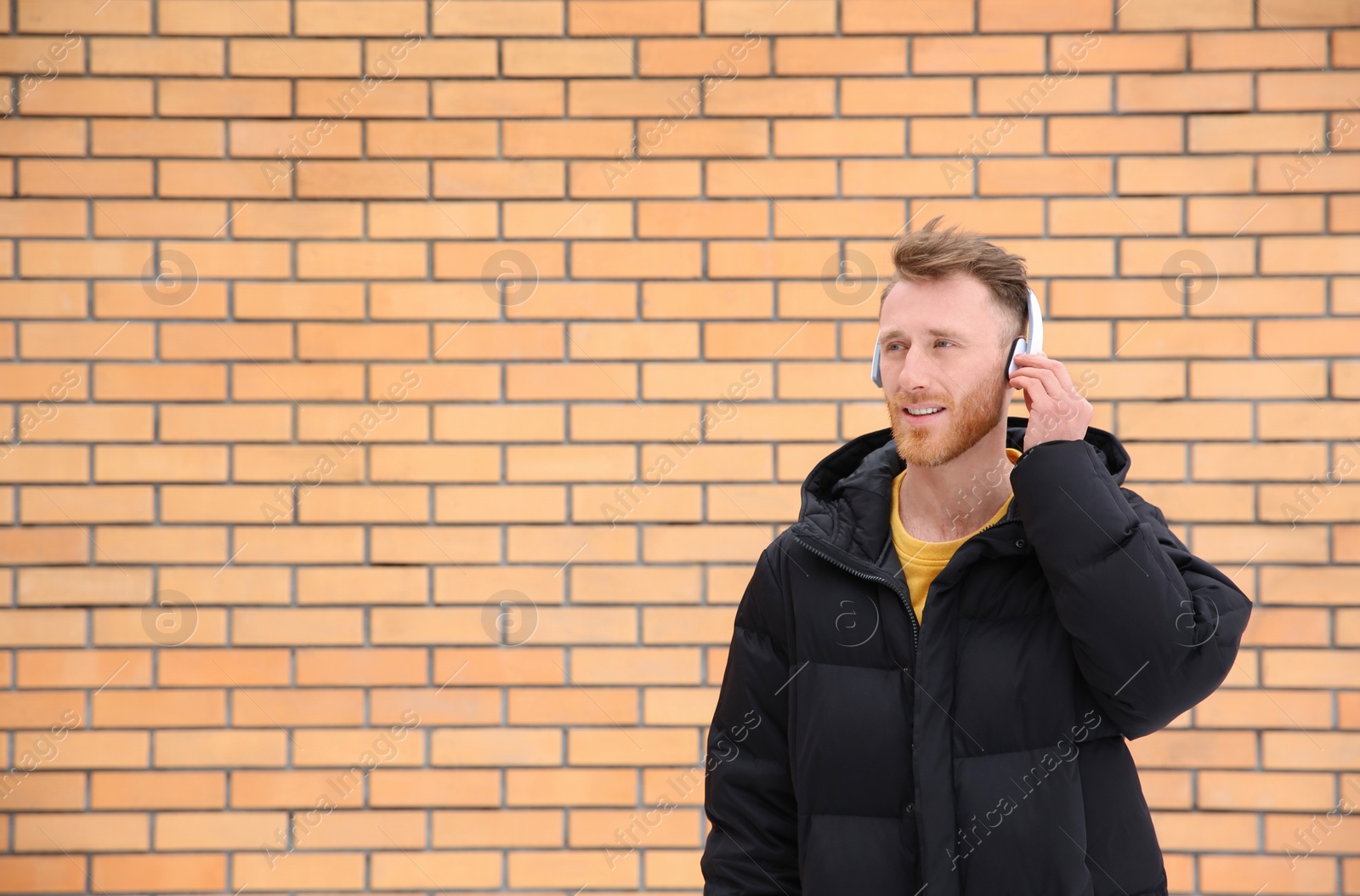 Photo of Young man listening to music with headphones against brick wall. Space for text