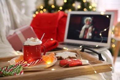 Photo of MYKOLAIV, UKRAINE - DECEMBER 25, 2020: Laptop displaying Christmas Chronicles movie, focus on tray with cocoa and snacks indoors. Cozy winter holidays atmosphere