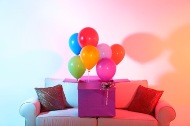 Gift box with bright air balloons on sofa against color background