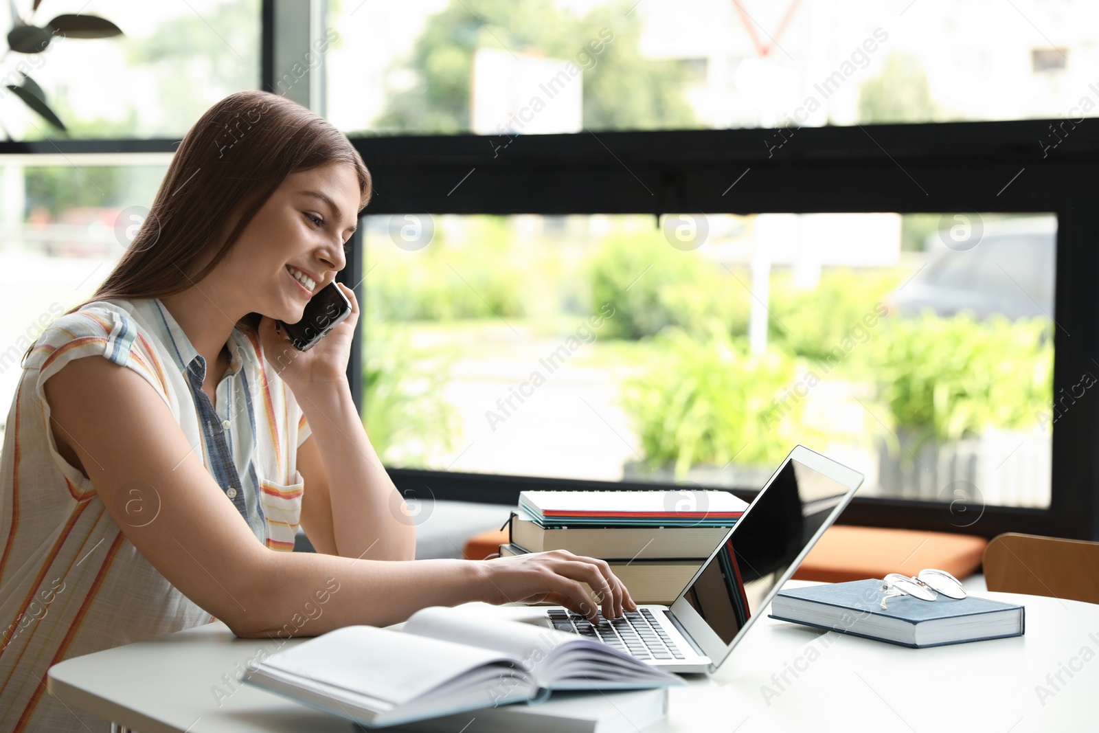 Photo of Young woman talking on phone and working with laptop at table in library