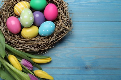 Photo of Colorful eggs in nest and tulips on blue wooden background, flat lay with space for text. Happy Easter