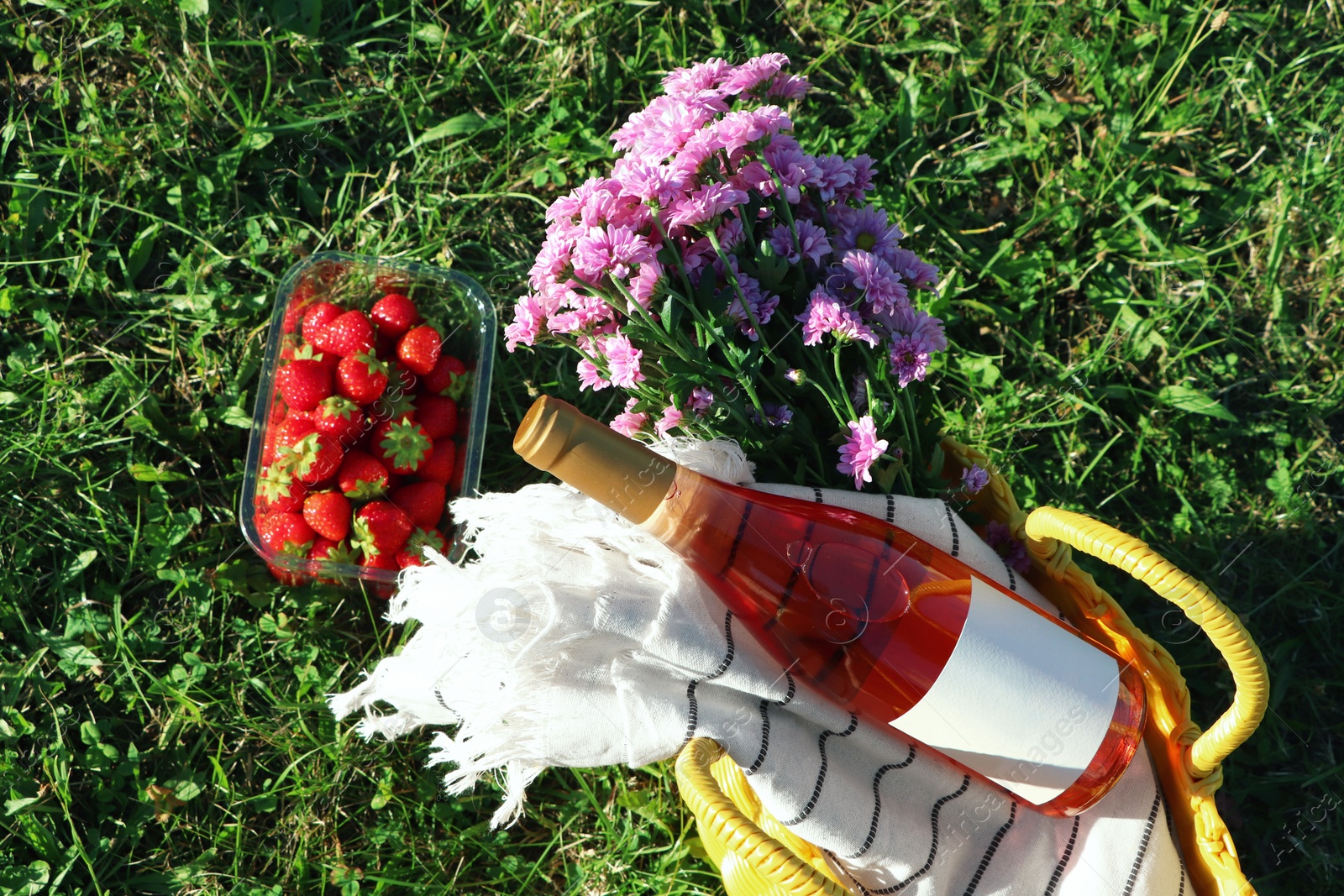 Photo of Yellow wicker bag with bottle of wine, strawberries, picnic blanket and beautiful flowers on green grass outdoors