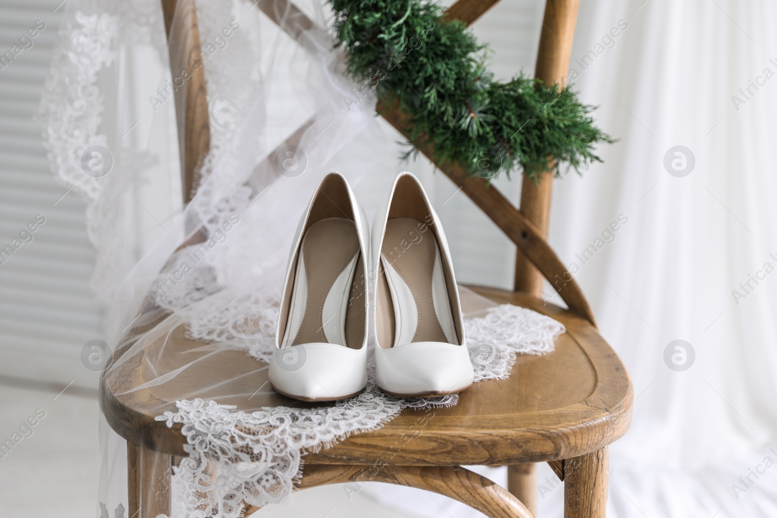 Photo of Pair of white high heel shoes, veil and wreath on wooden chair indoors. Dressing for wedding