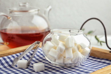 Photo of Refined sugar cubes in glass bowl and tongs on wooden tray