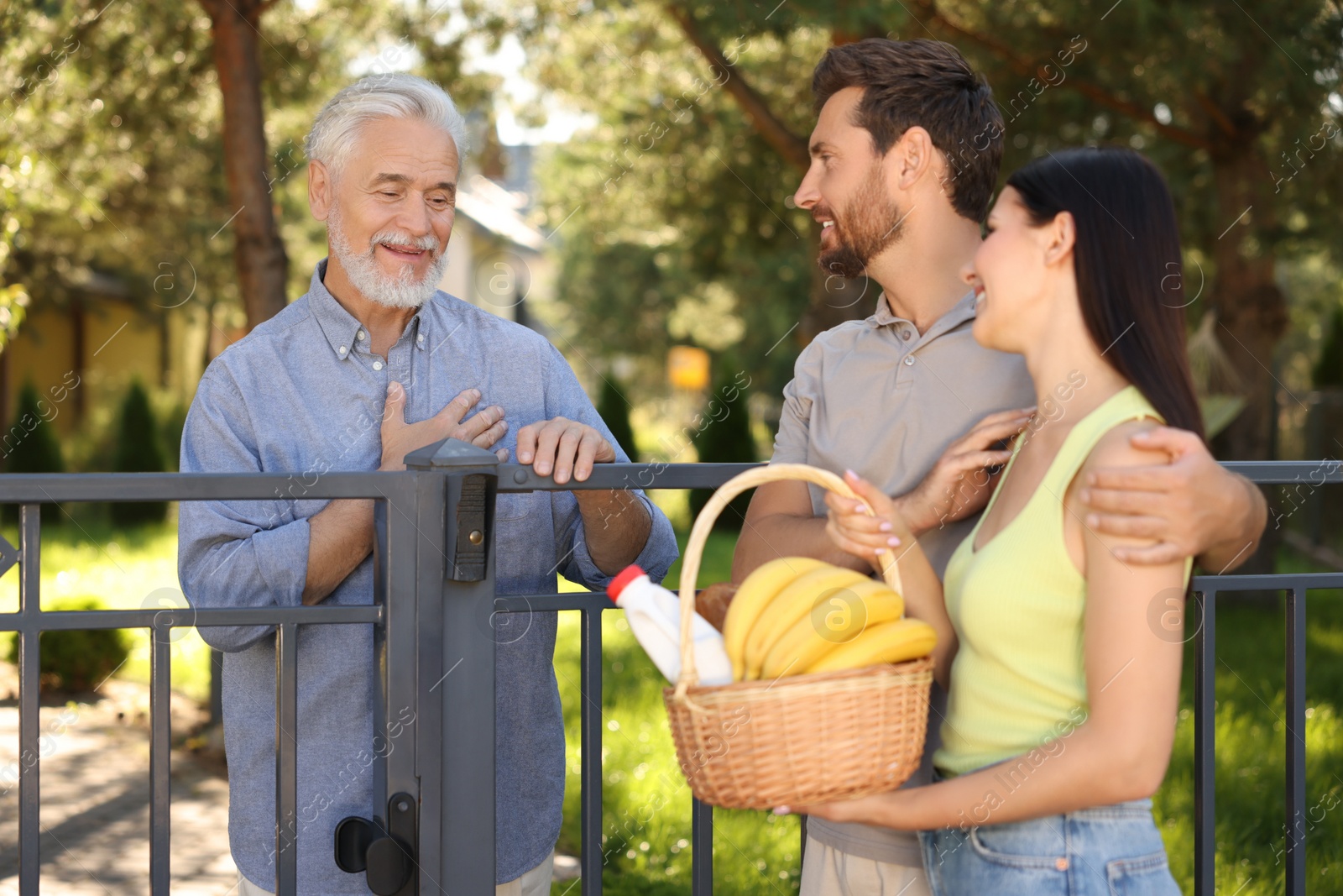 Photo of Friendly relationship with neighbours. Young couple with wicker basket of products treating senior man near fence outdoors