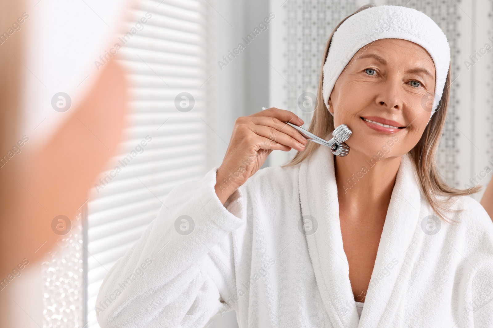 Photo of Woman massaging her face with metal roller near mirror in bathroom