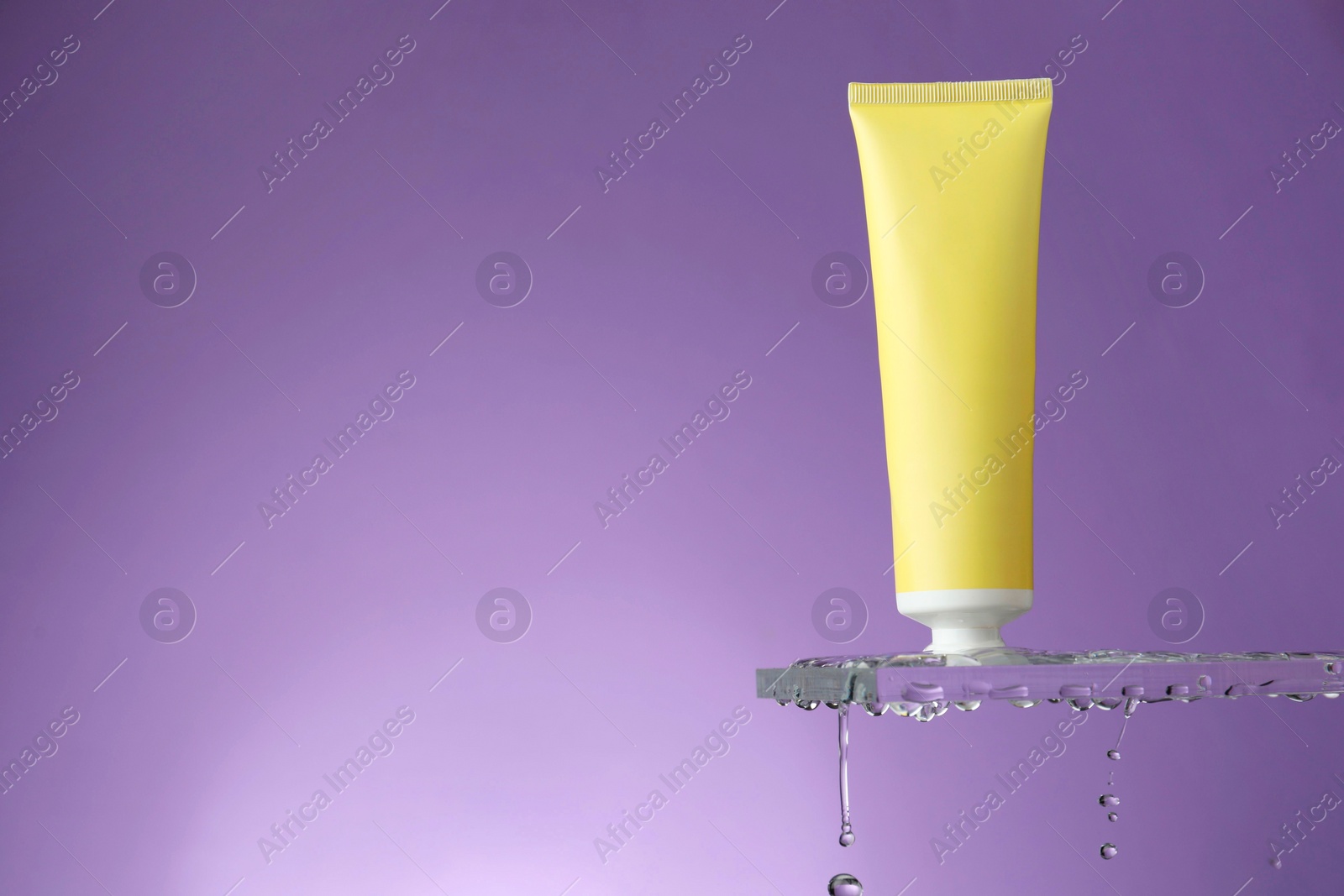 Photo of Moisturizing cream in tube on glass with water drops against violet background. Space for text