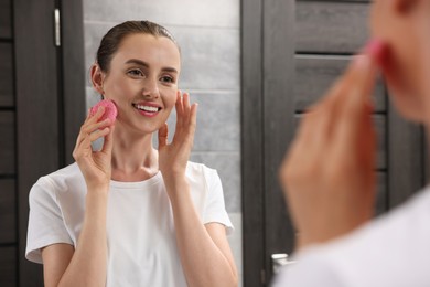 Happy young woman washing her face with sponge near mirror in bathroom