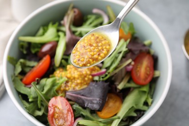 Photo of Pouring tasty vinegar based sauce (Vinaigrette) from spoon into bowl with salad at grey table, closeup