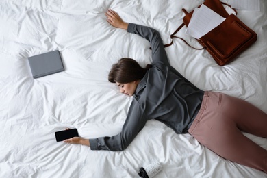 Photo of Exhausted woman with smartphone and documents sleeping fully dressed on bed at home, above view