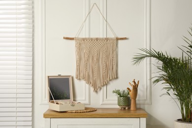 Photo of Open jewelry box and decor elements on chest of drawers near white wall with stylish macrame