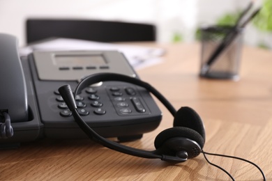 Photo of Desktop telephone and headset on wooden table in office, closeup. Hotline service