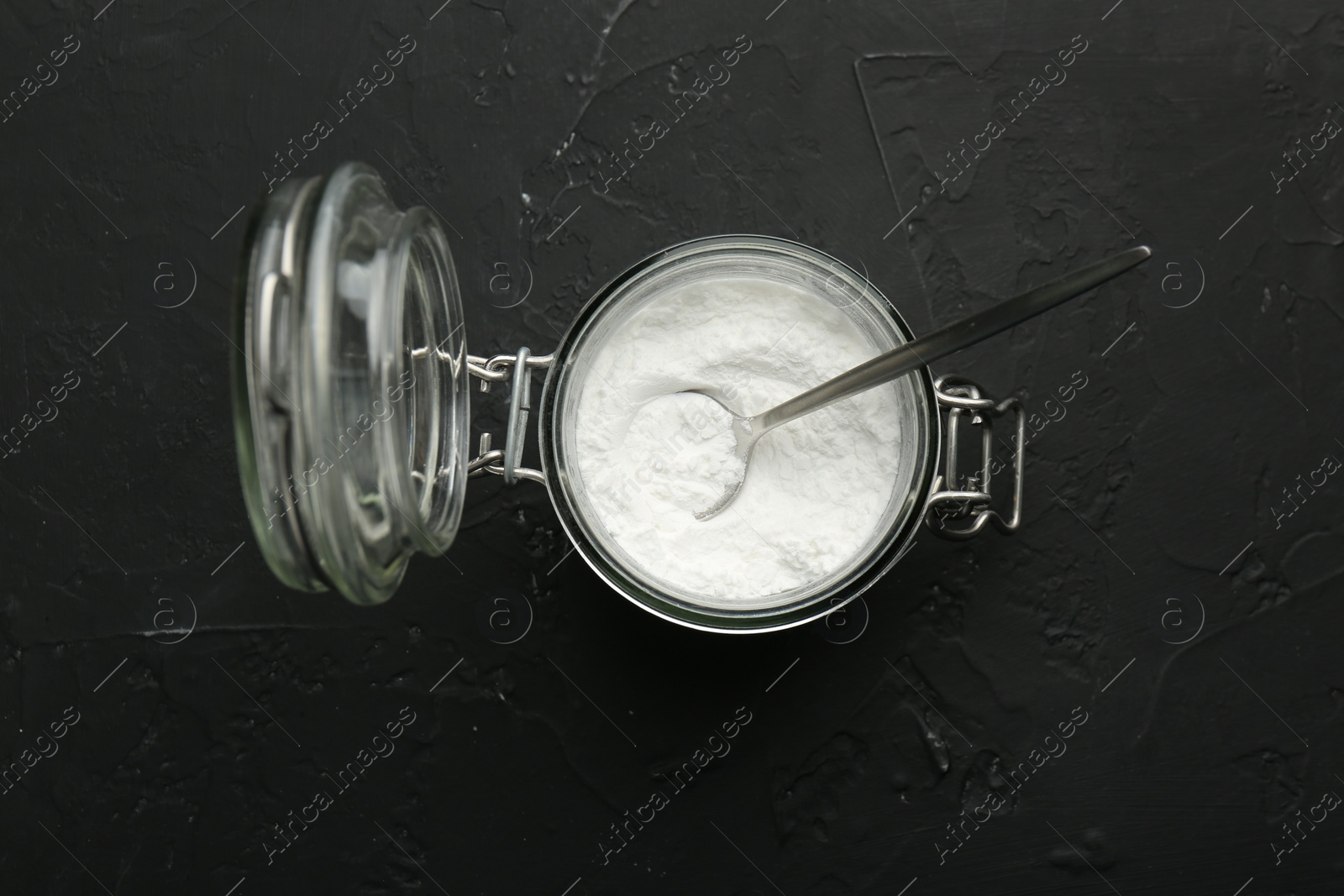 Photo of Baking powder in jar and spoon on black textured table, top view