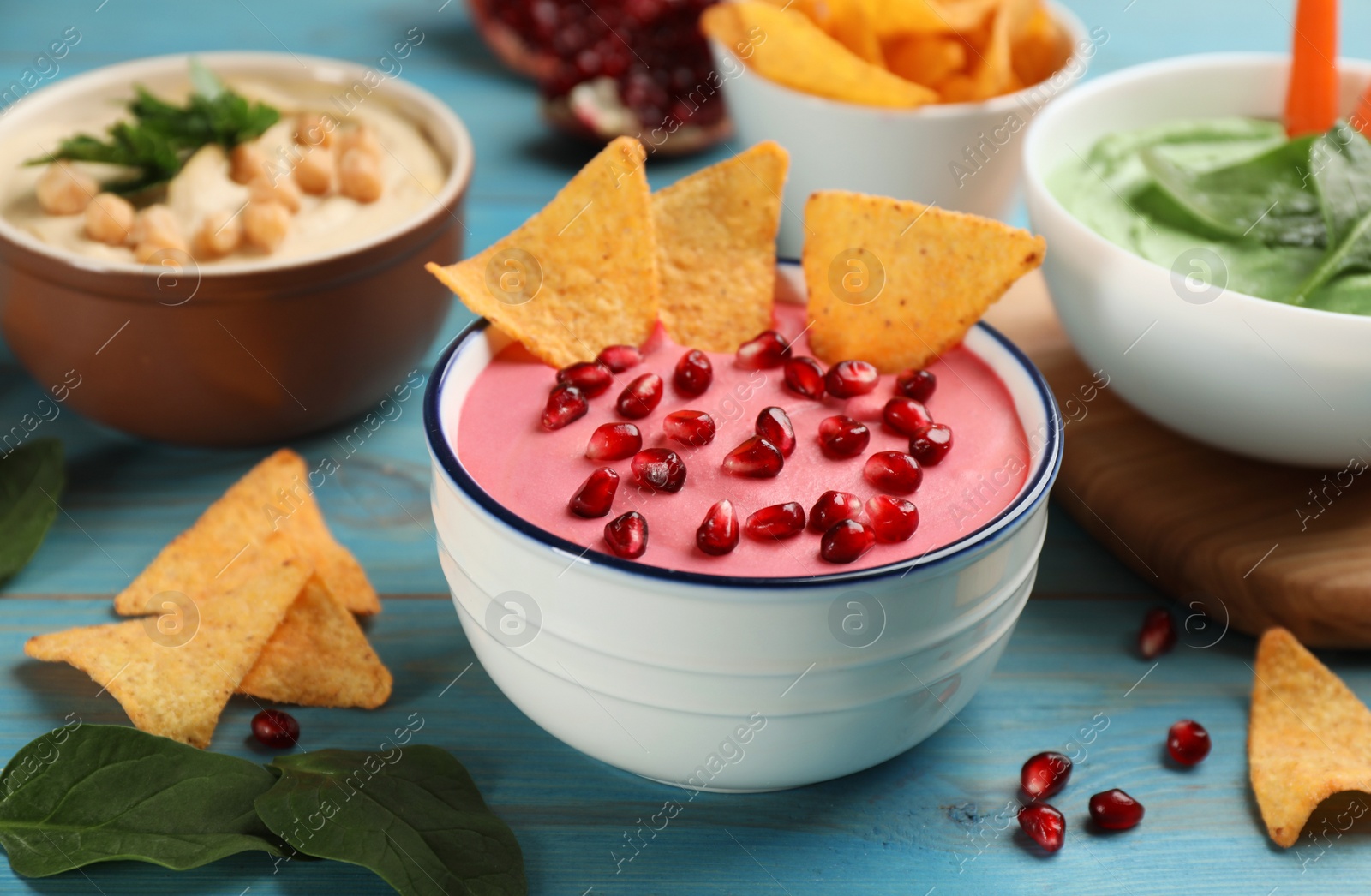 Photo of Bowl of delicious pink hummus with tortilla chips and pomegranate seeds on light blue wooden table