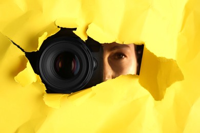 Photo of Hidden woman with camera spying through torn hole in yellow paper, closeup