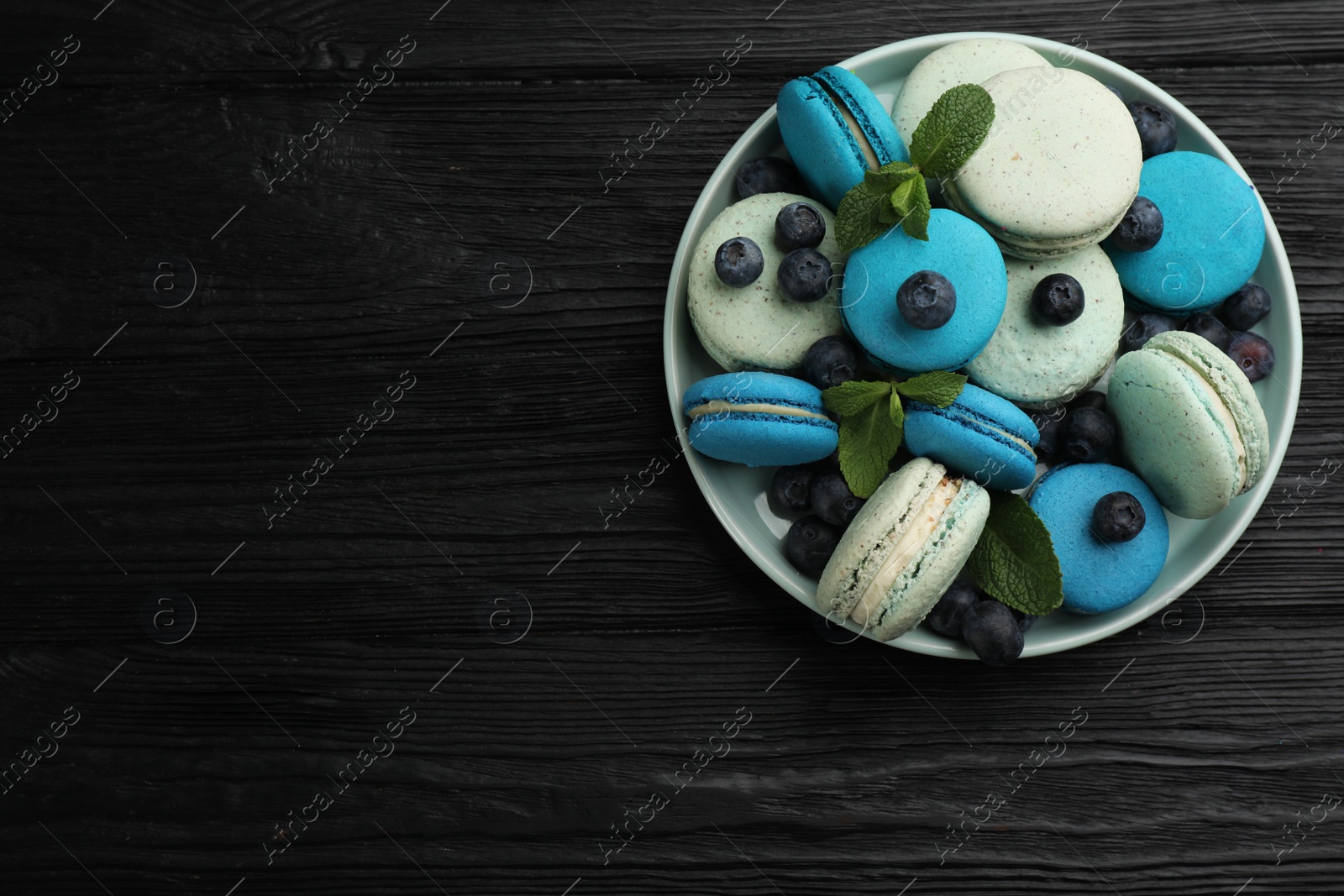 Photo of Delicious macarons, mint and blueberries in bowl on black wooden table, top view. Space for text